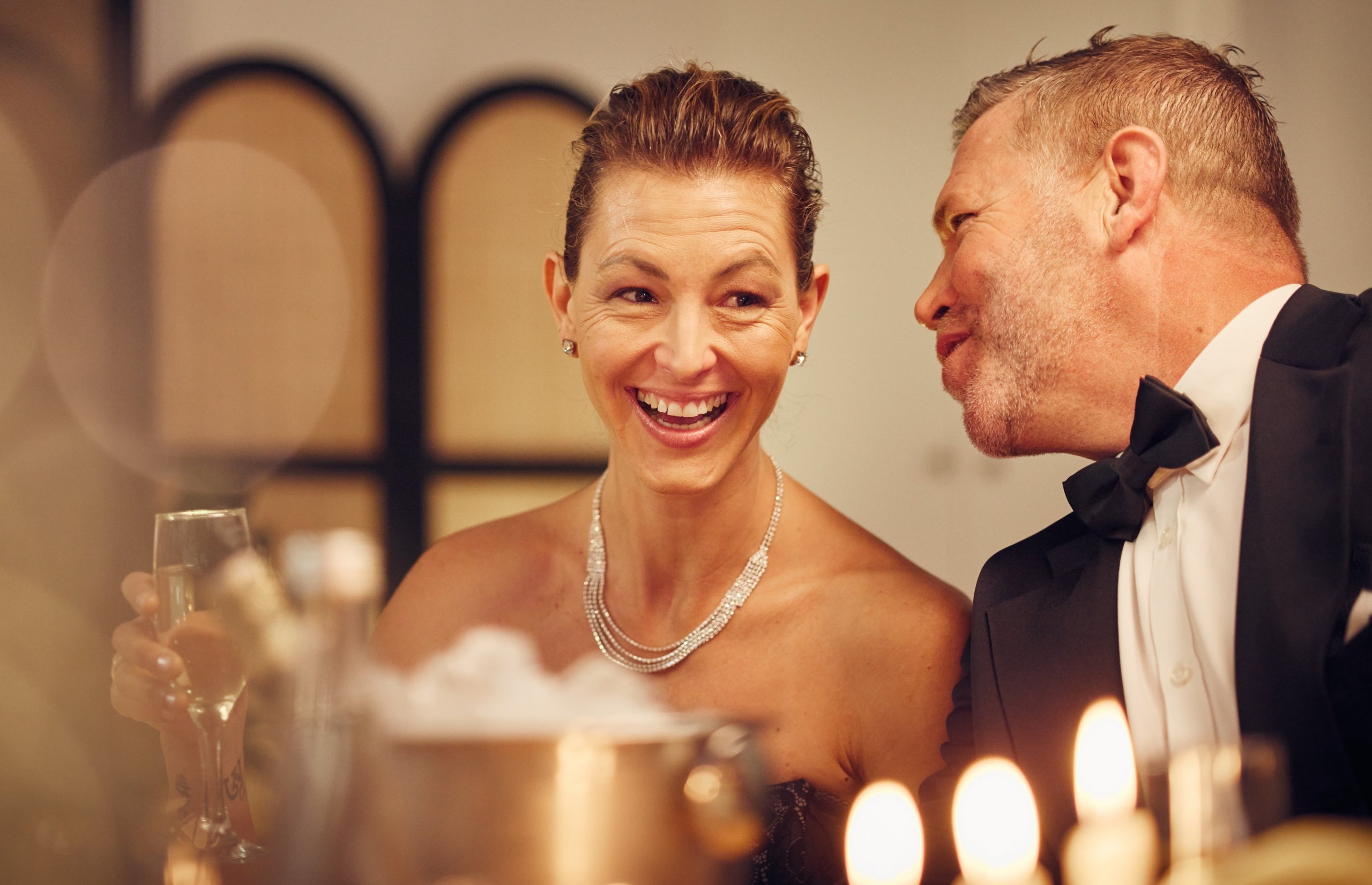 A man whispers something into a smiling woman's ear at a social event