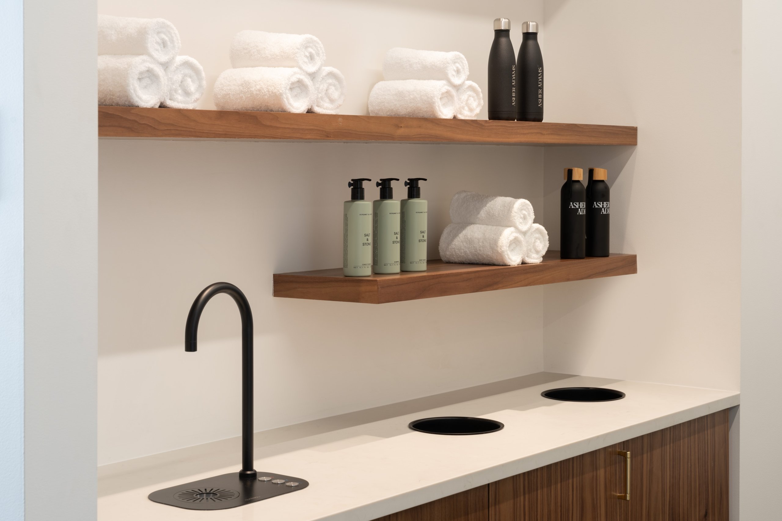 Wooden shelves holding clear towels, reusable water bottles and Salt & Stone soap above a filtered water station at the fitness facility in Asher Adams Hotel, Salt Lake City