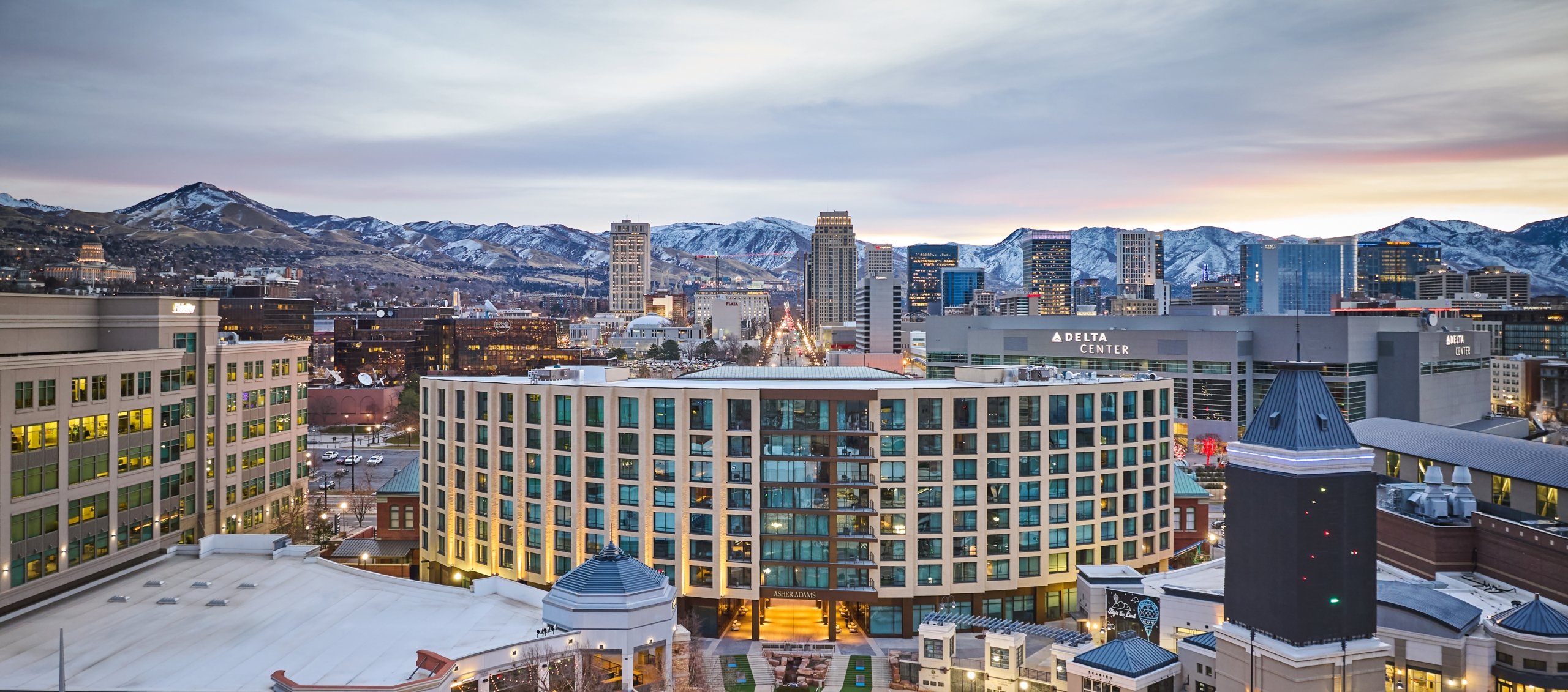 The exterior of Asher Adams hotel, the Delta Center, and Salt Lake City's snow-capped mountains